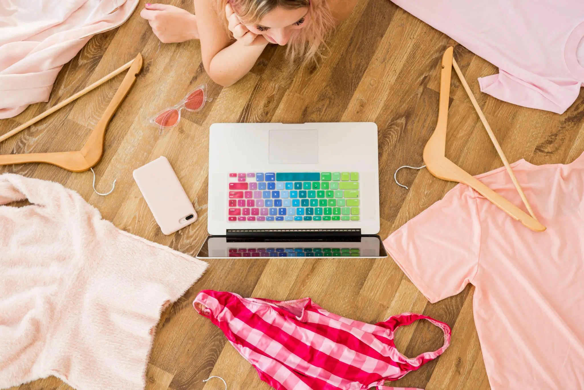 Person lying on wooden floor with laptop, pink clothing, and accessories scattered around