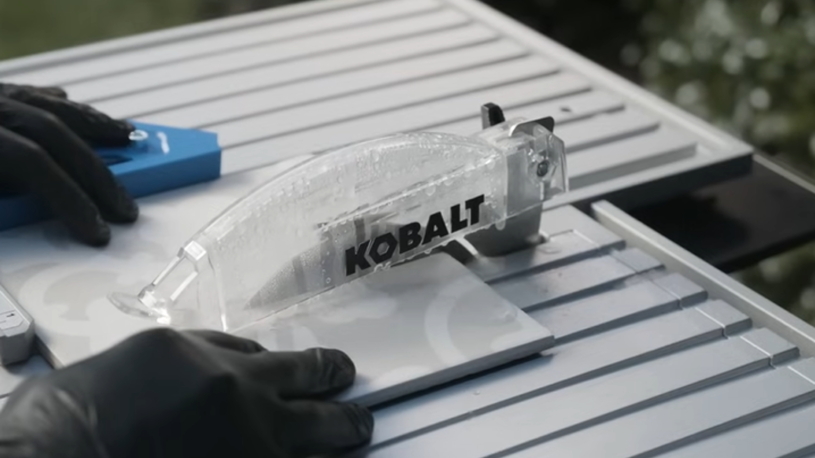 Gloved hands guiding a tile through a wet tile saw with a clear blade guard on a worktable
