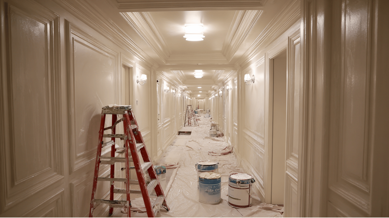 Hallway with freshly painted walls, tall ladders, ceiling rollers, and painting supplies indoors neatly.