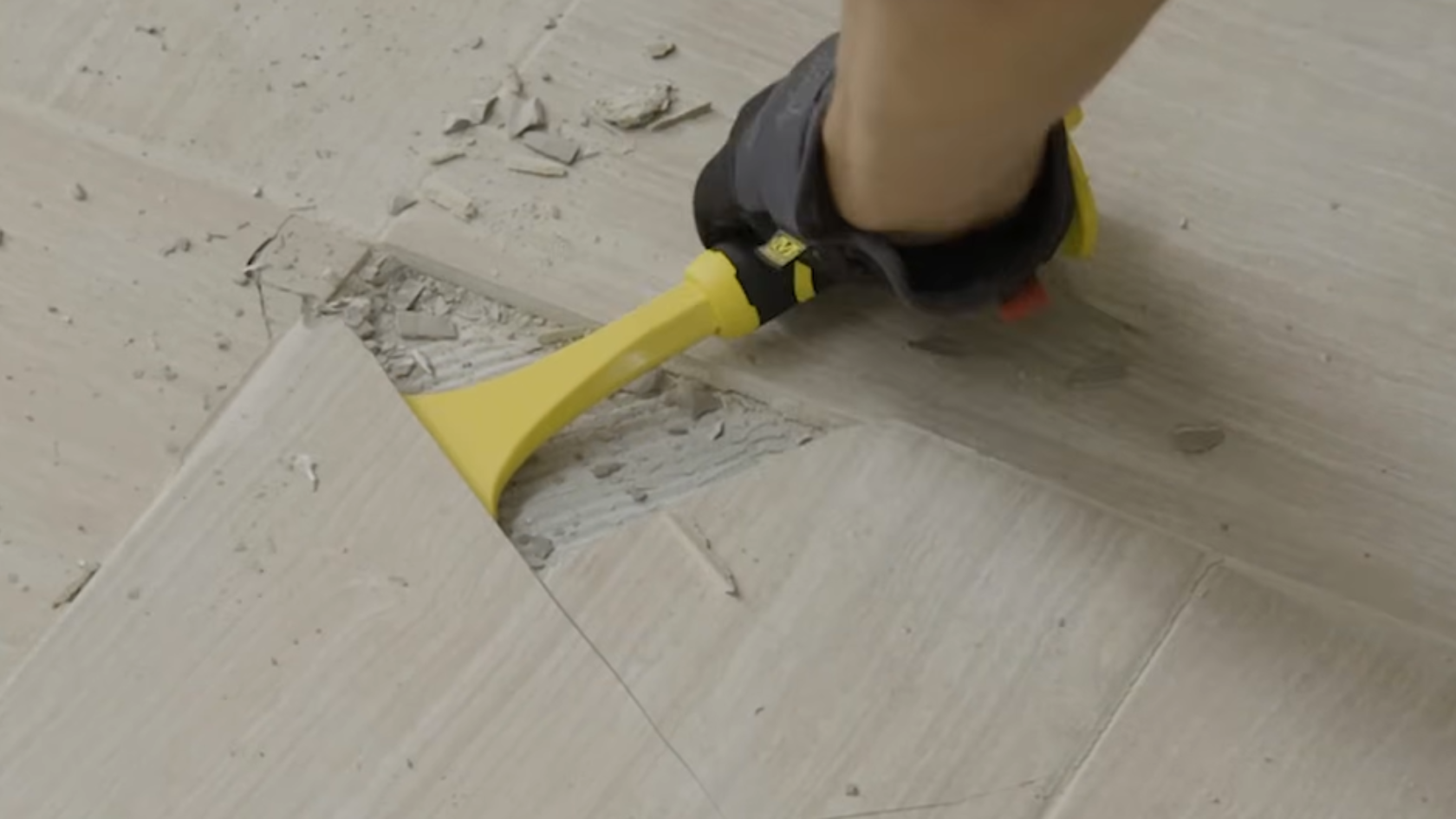 Hand using a yellow scraper tool to remove flooring, lifting a tile and exposing concrete and debris underneath