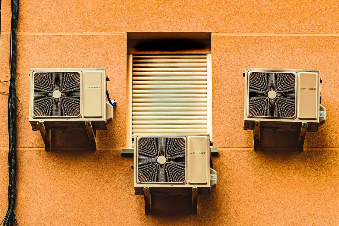 Three air conditioning units mounted on orange exterior wall near a covered window