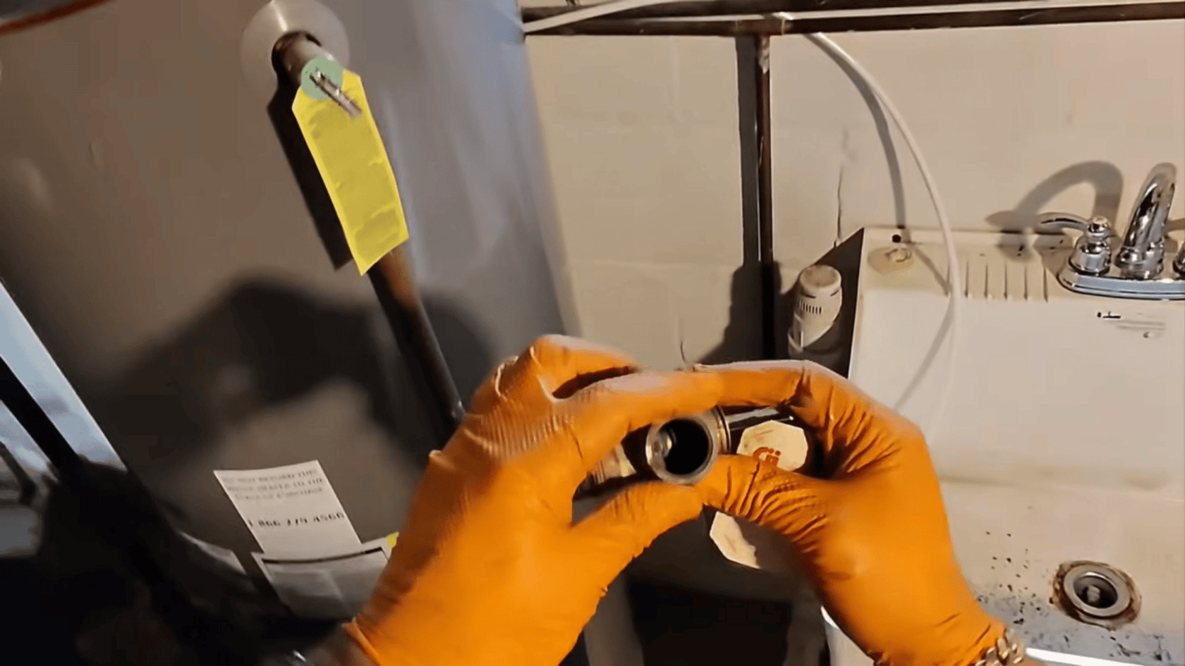 Hands wearing orange gloves holding a small boiler part, with a water heater and pipes in the background.