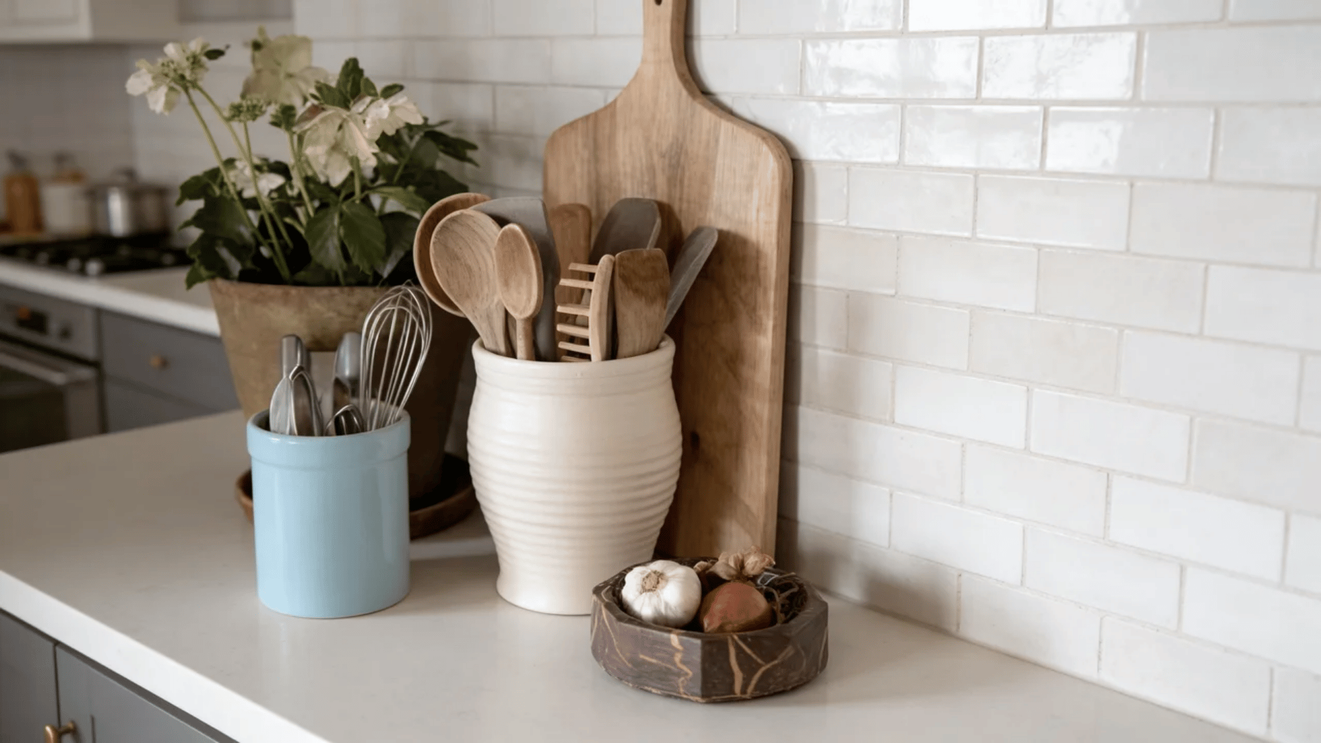 Kitchen countertop with wooden utensils, cutting board, flowers, and white tile backsplash.