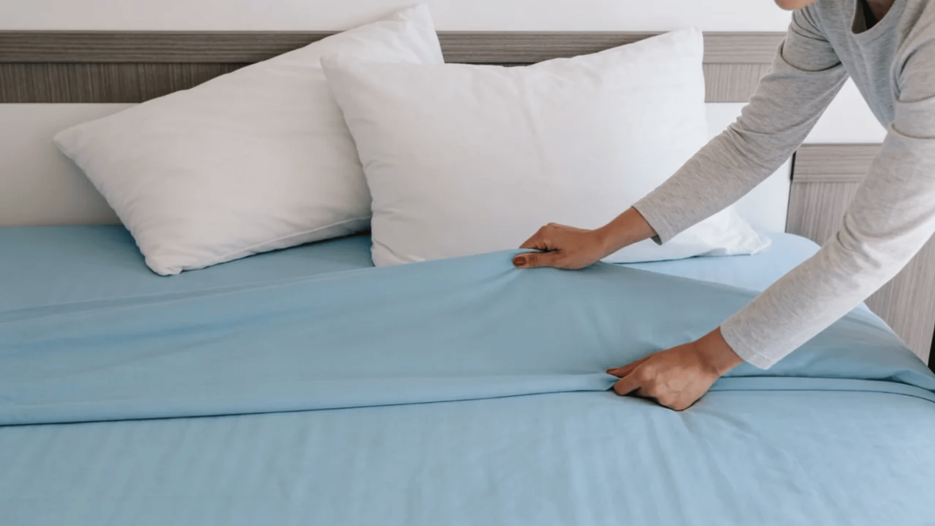 Person making a bed with light blue sheets and white pillows in a bedroom.