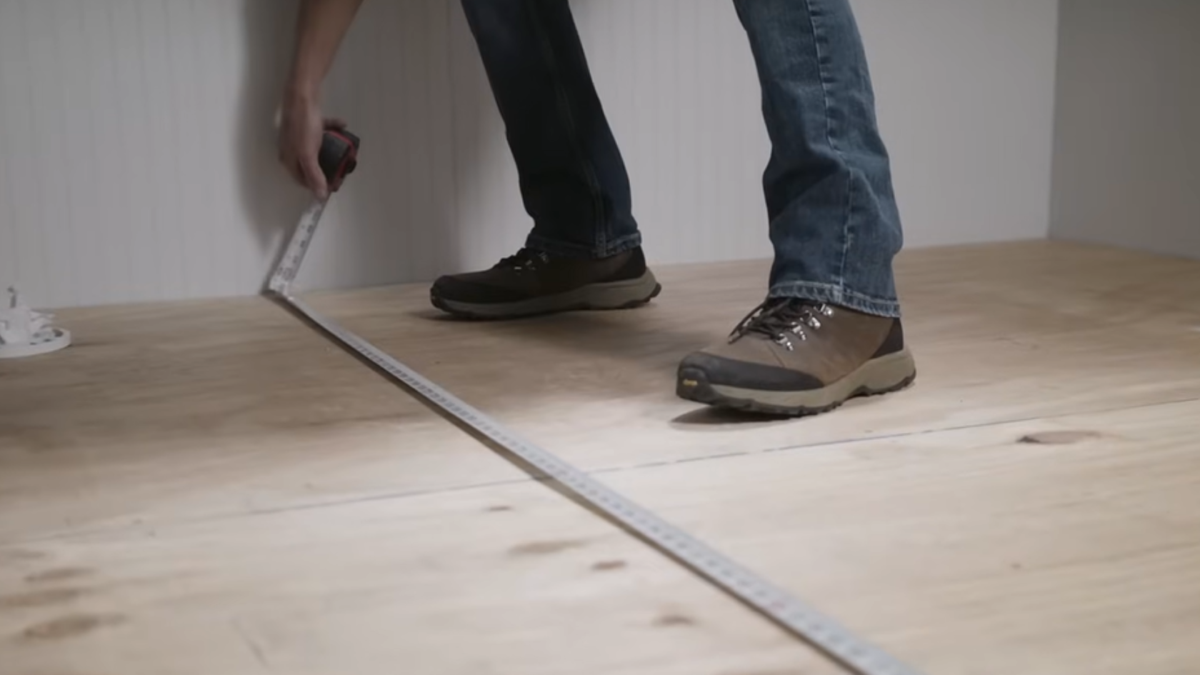 Person measuring a floor with a tape measure, standing on plywood subfloor in a room under construction
