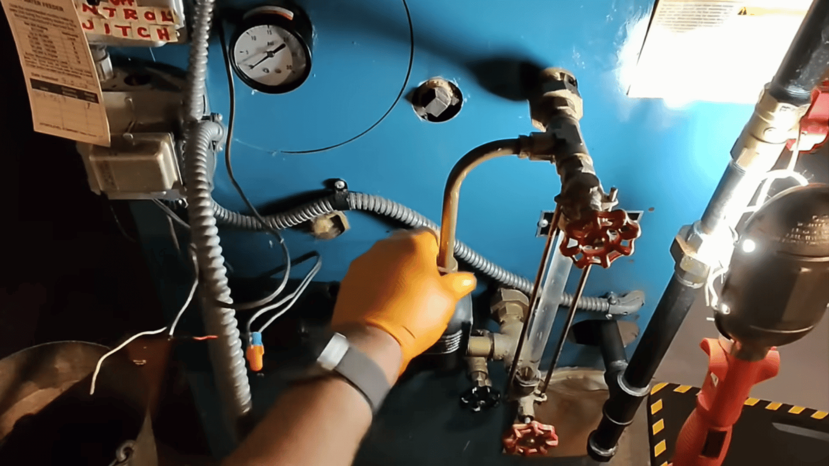 Hand wearing an orange glove adjusting a valve on a boiler, with gauges and pipes visible in the background.