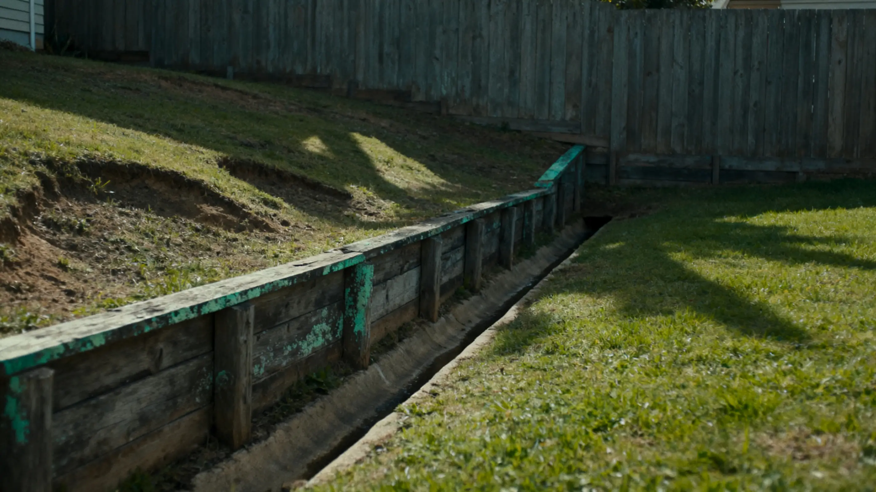 Wooden retaining wall and drainage channel on sloped grassy backyard with wooden fence