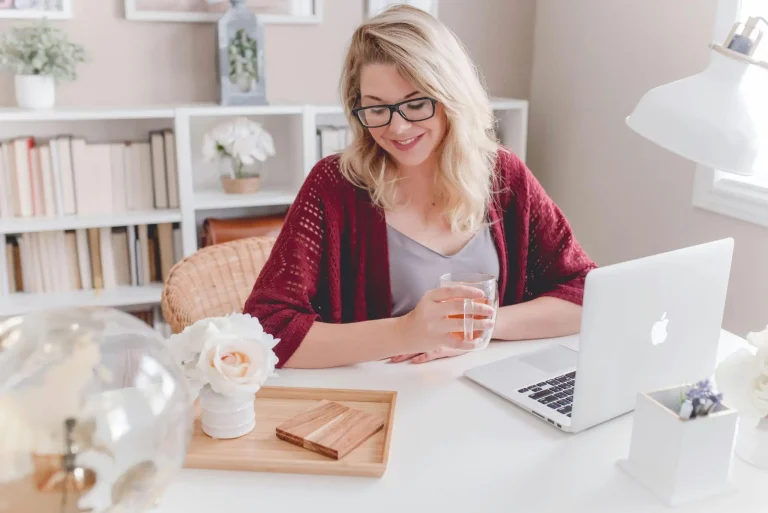 Woman with glasses holding cup at laptop in bright home office setting