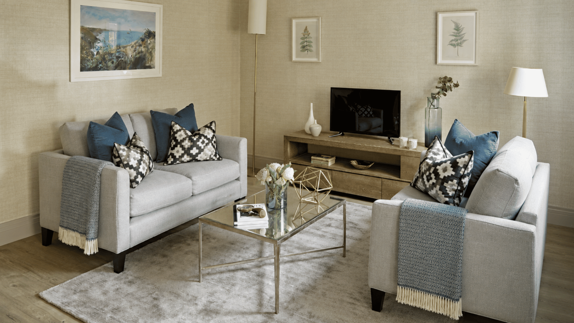 Two gray loveseats with blue pillows, a glass coffee table, and TV in a small living room.
