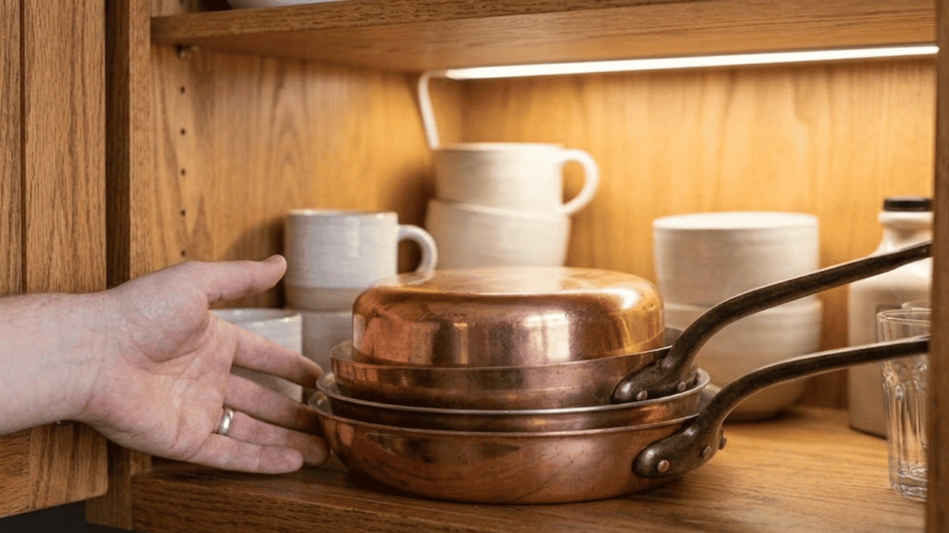 a hand reaching toward a stack of copper pans inside a lit wooden cabinet