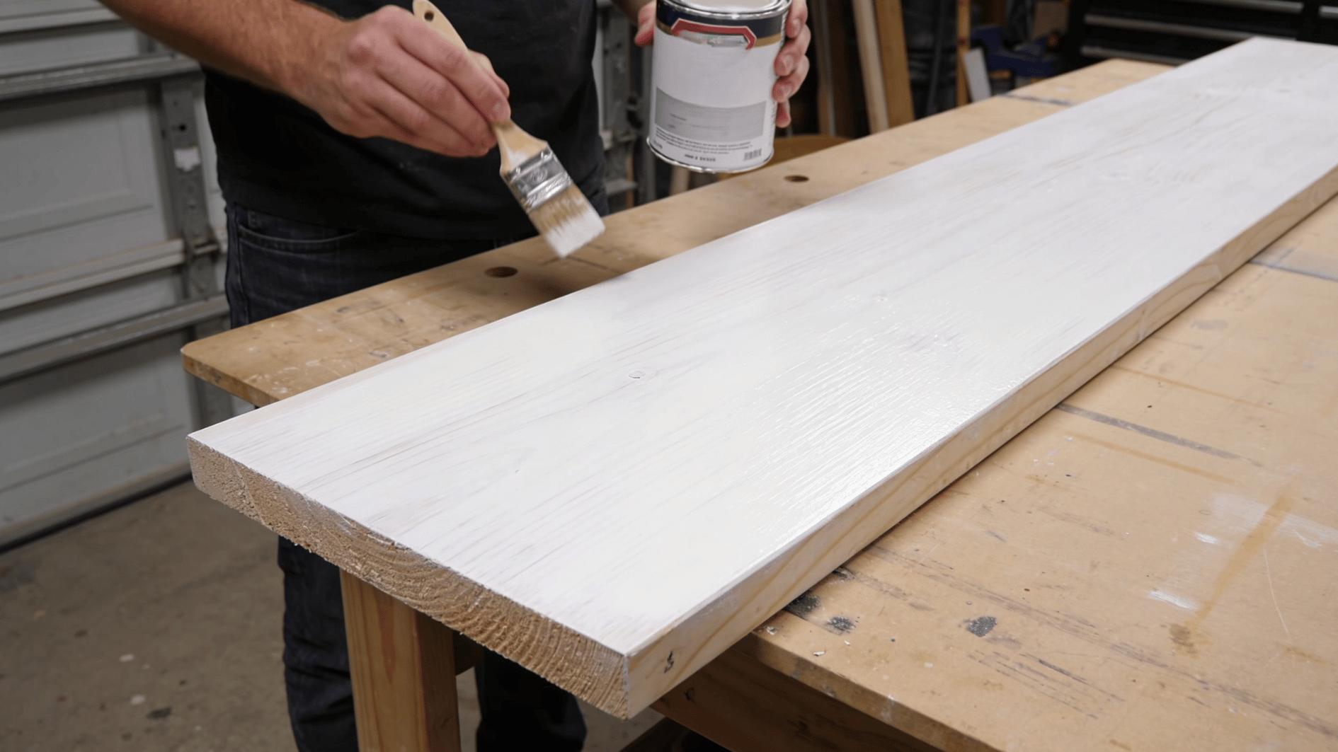 A person steps back slightly, one hand holding a can of matte water-based sealer and a brush, assessing the even milky-white finish.