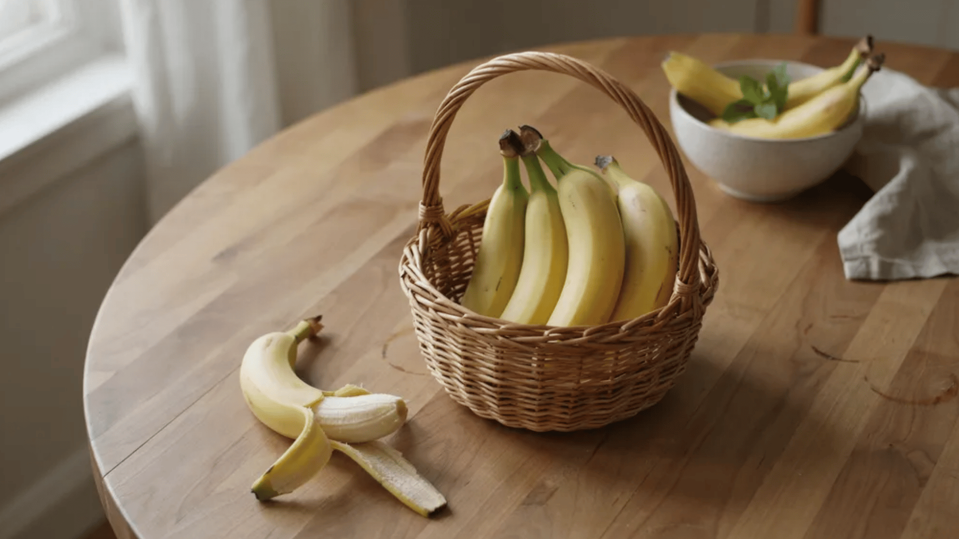 basket of ripe bananas on a wooden table with a partially peeled banana and a bowl of bananas in the background