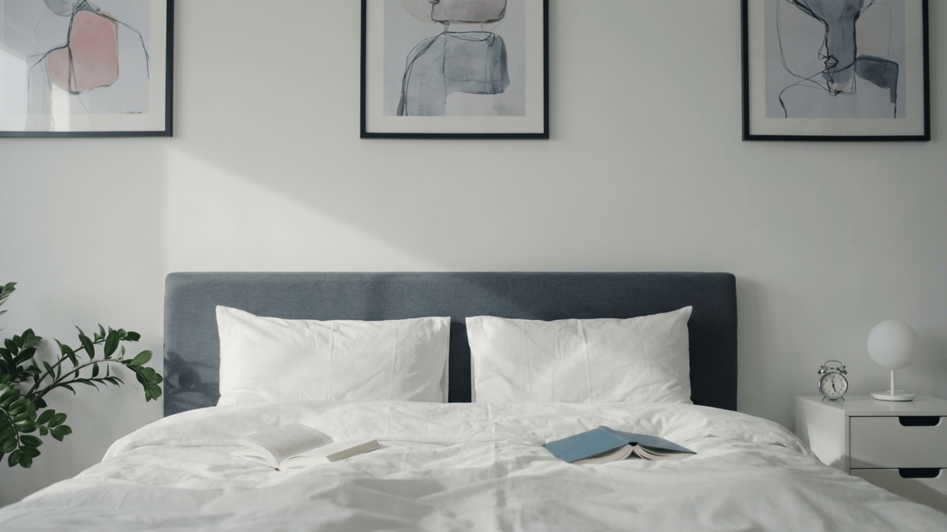 Minimalist guest bed with crisp white bedding, a gray headboard, framed artwork, and open books on the mattress