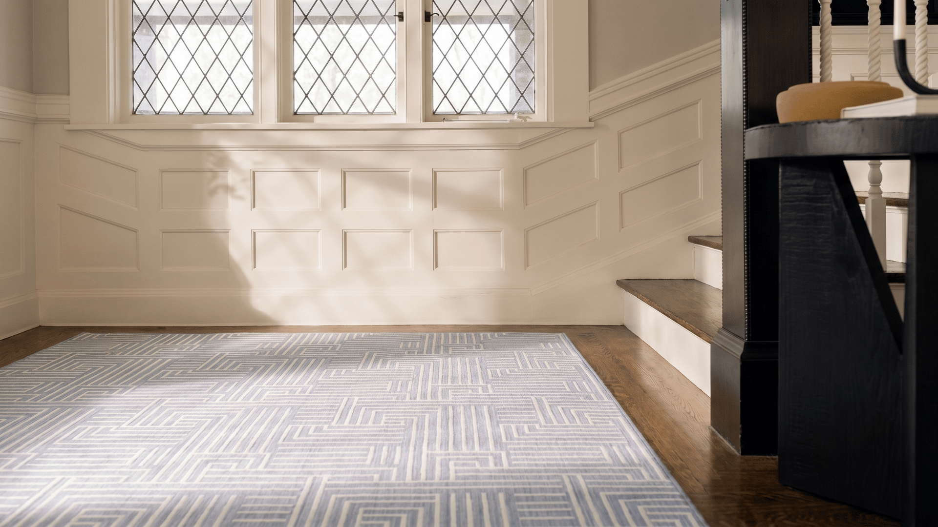A light blue geometric patterned Ernesta rug styled in a classic entryway with hardwood floors and white wainscoting paneling.