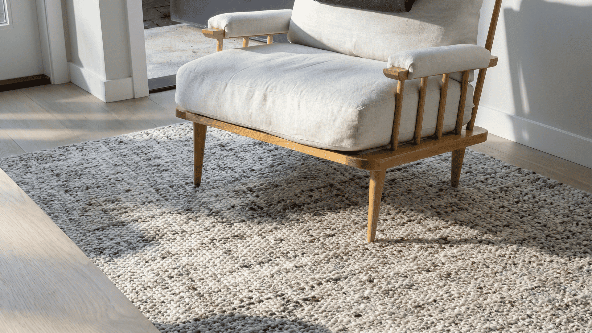 A chunky wool Ernesta rug in mixed grey and cream tones styled under a mid-century wood lounge chair in a bright, minimal living space.