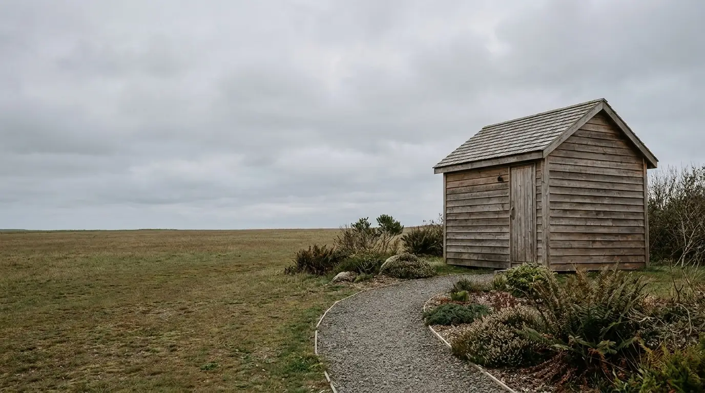 Wooden shed on a gravel path surrounded by grass and shrubs under cloudy sky