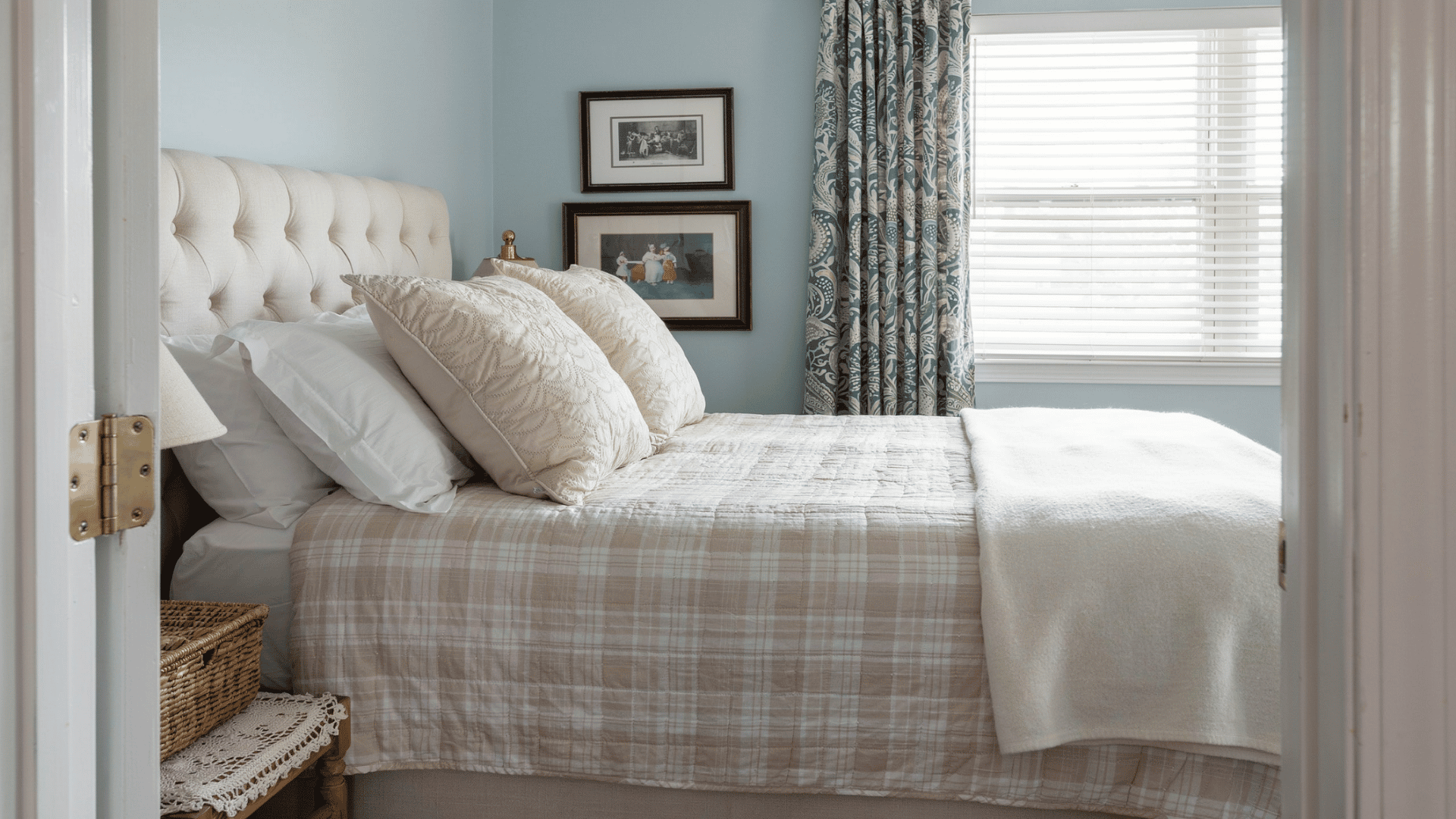 Cozy guest room with a tufted headboard, plaid bedding, patterned curtains, and a window with blinds