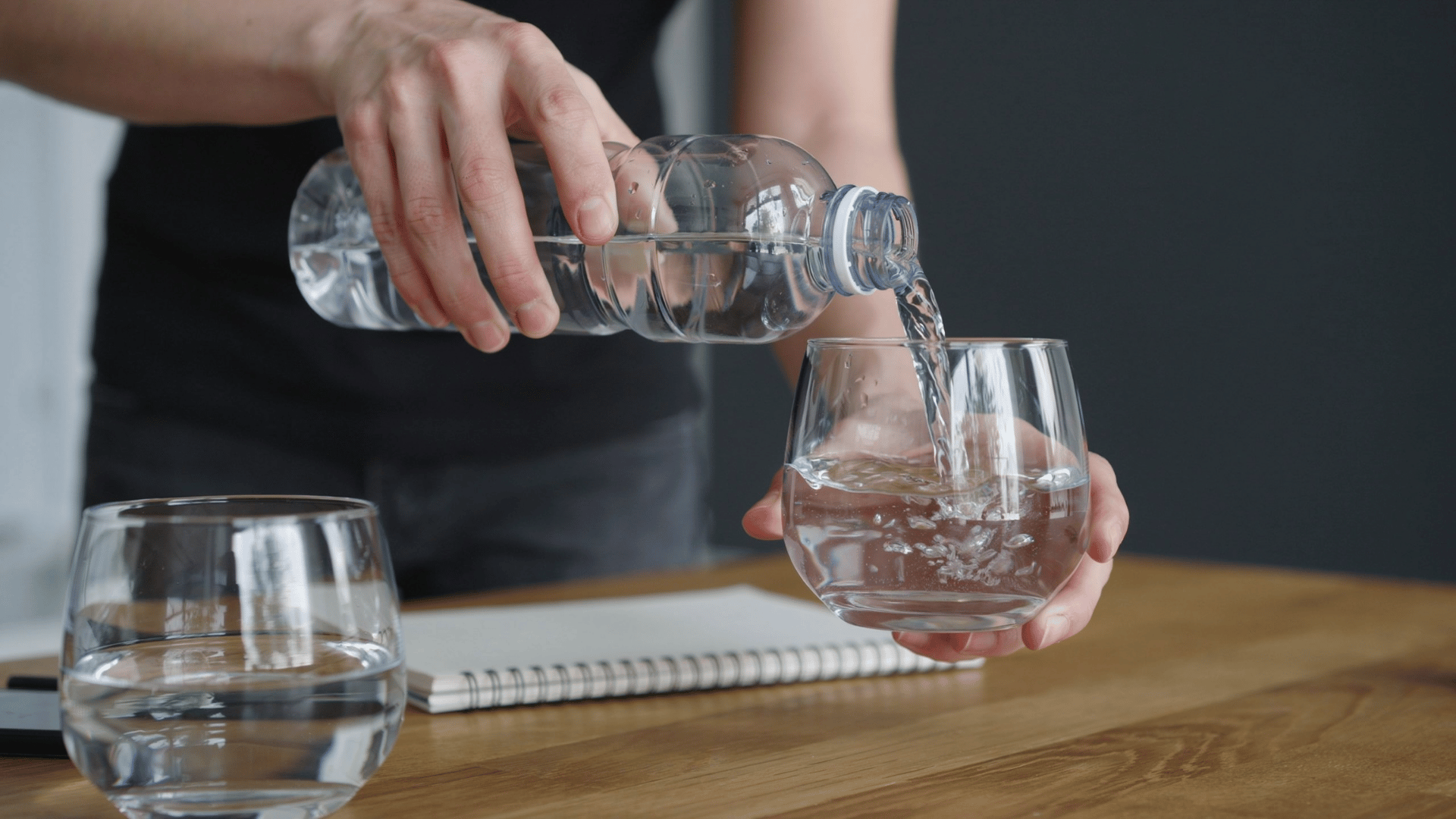 A person pours water from a clear plastic bottle into a glass on a wooden desk with a notebook, ensuring guest room hydration