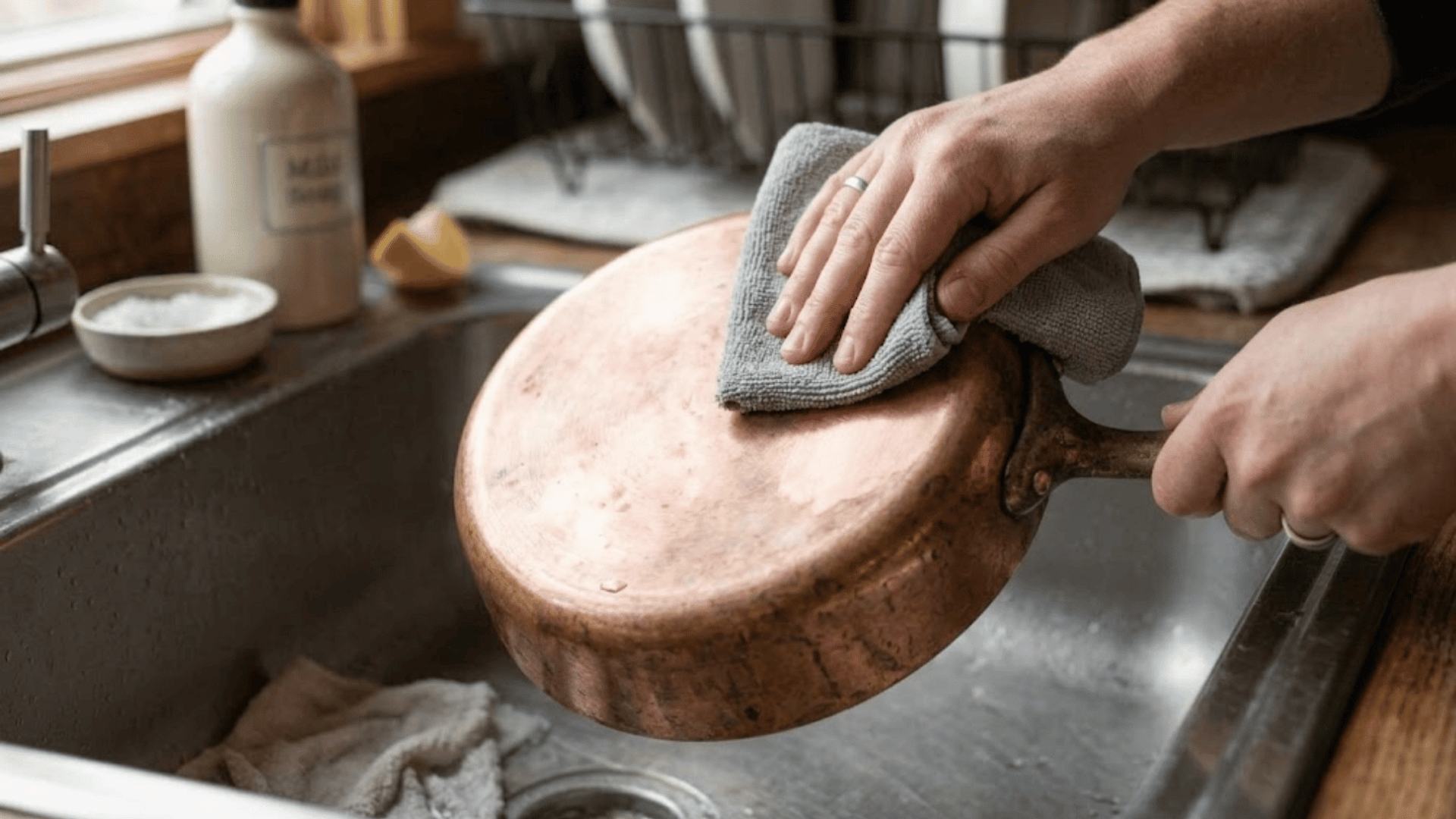 hands drying the bottom of a copper pan with a grey cloth over a kitchen sink