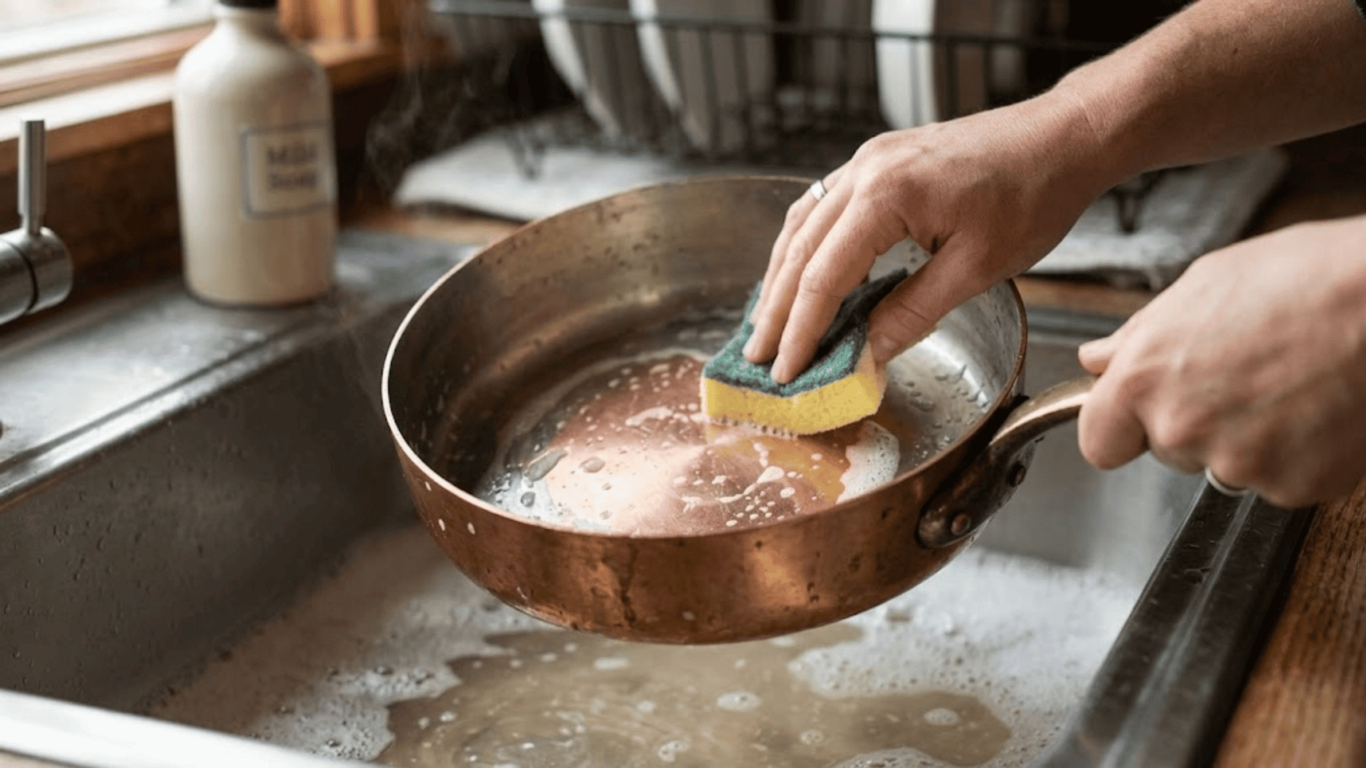 hands scrubbing a copper pan with a sponge in a soapy sink