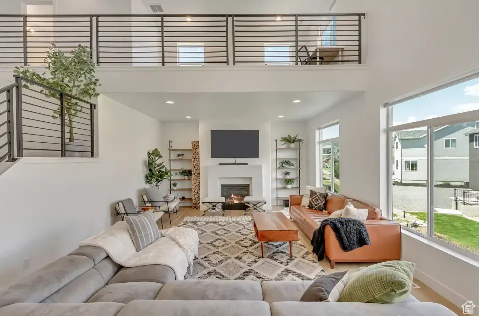 Spacious living room with modern furniture, fireplace, large windows, and lofted second floor railing