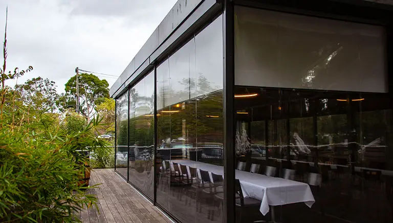 Glass-walled dining area with long table set for guests, surrounded by greenery