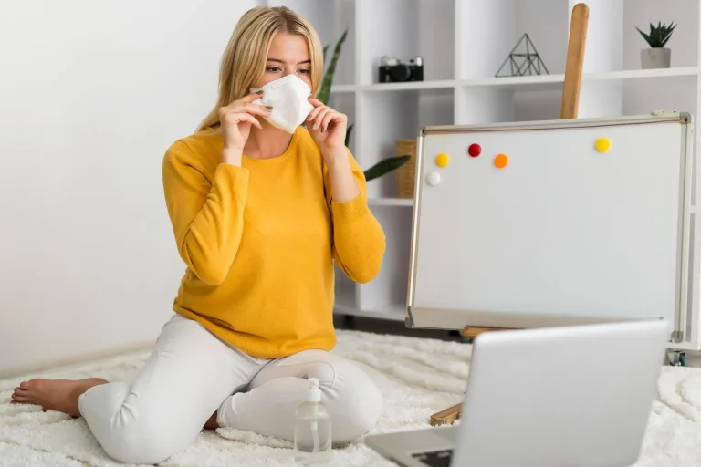 Woman in yellow sweater applying face mask, sitting on white rug near laptop and whiteboard