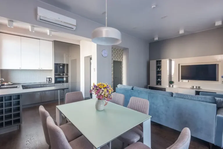Modern living room and kitchen with blue sofa, dining table, and wall-mounted TV in neutral tones