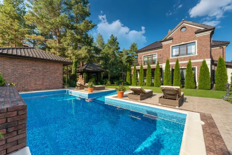 Outdoor swimming pool with lounge chairs and brick house surrounded by trees under blue sky
