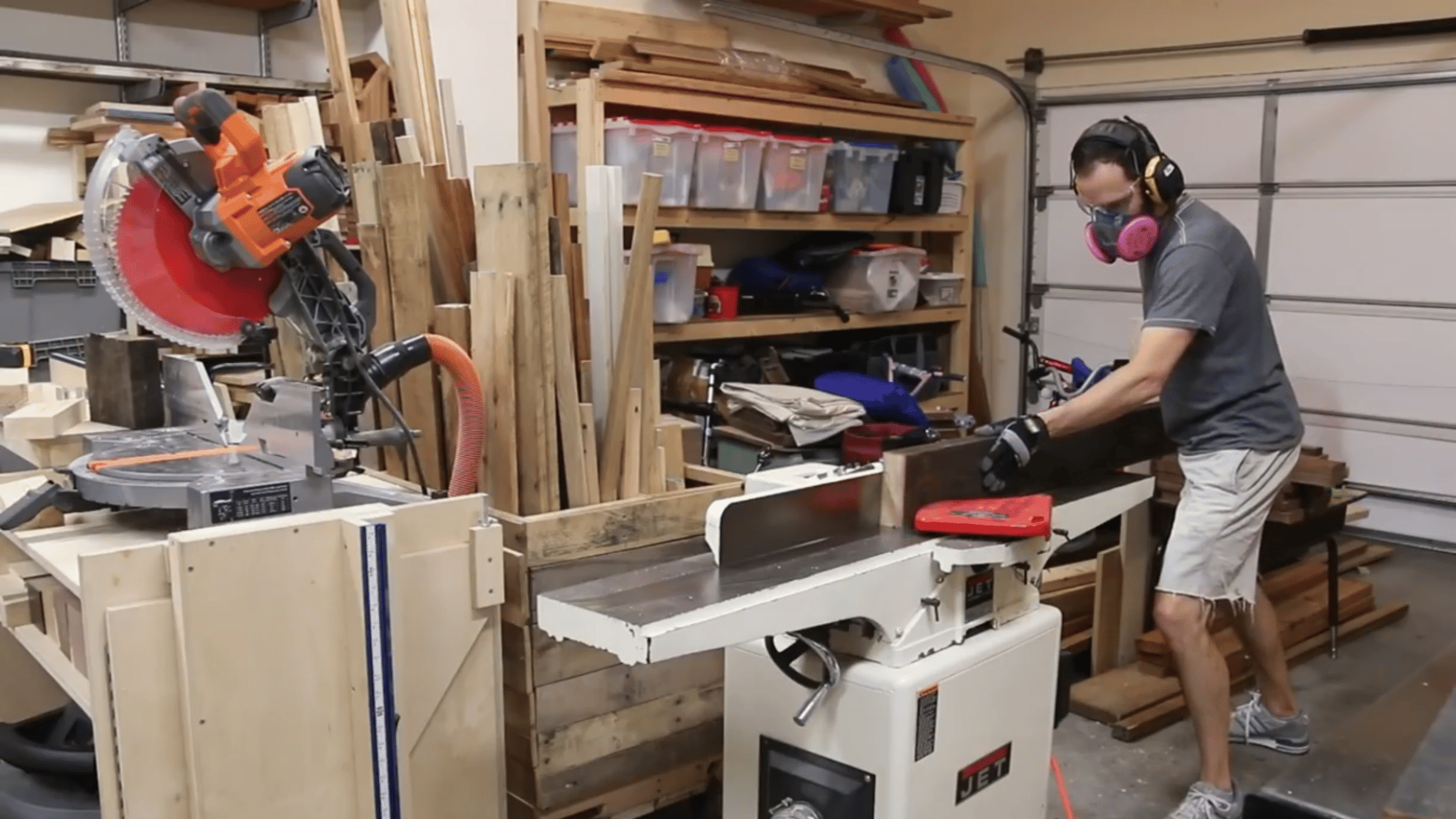 man smoothing a wooden tabletop board on a jointer while wearing protective gear in a small workshop