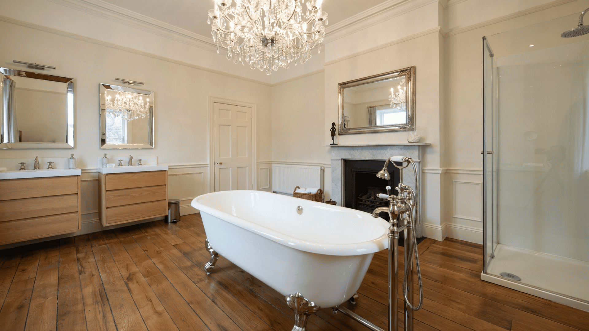 Elegant modern Victorian bathroom featuring a central clawfoot tub, crystal chandelier, double vanities, and a fireplace mantel