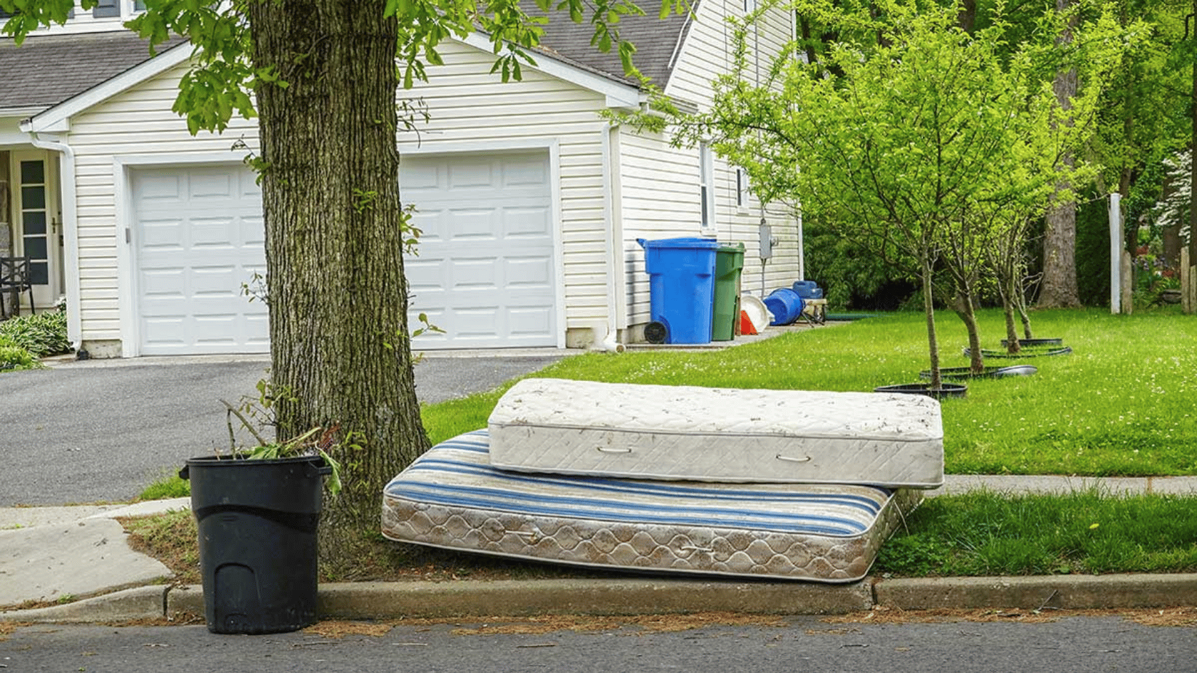 Two old mattresses left on curbside in a suburban driveway near a garage and trees