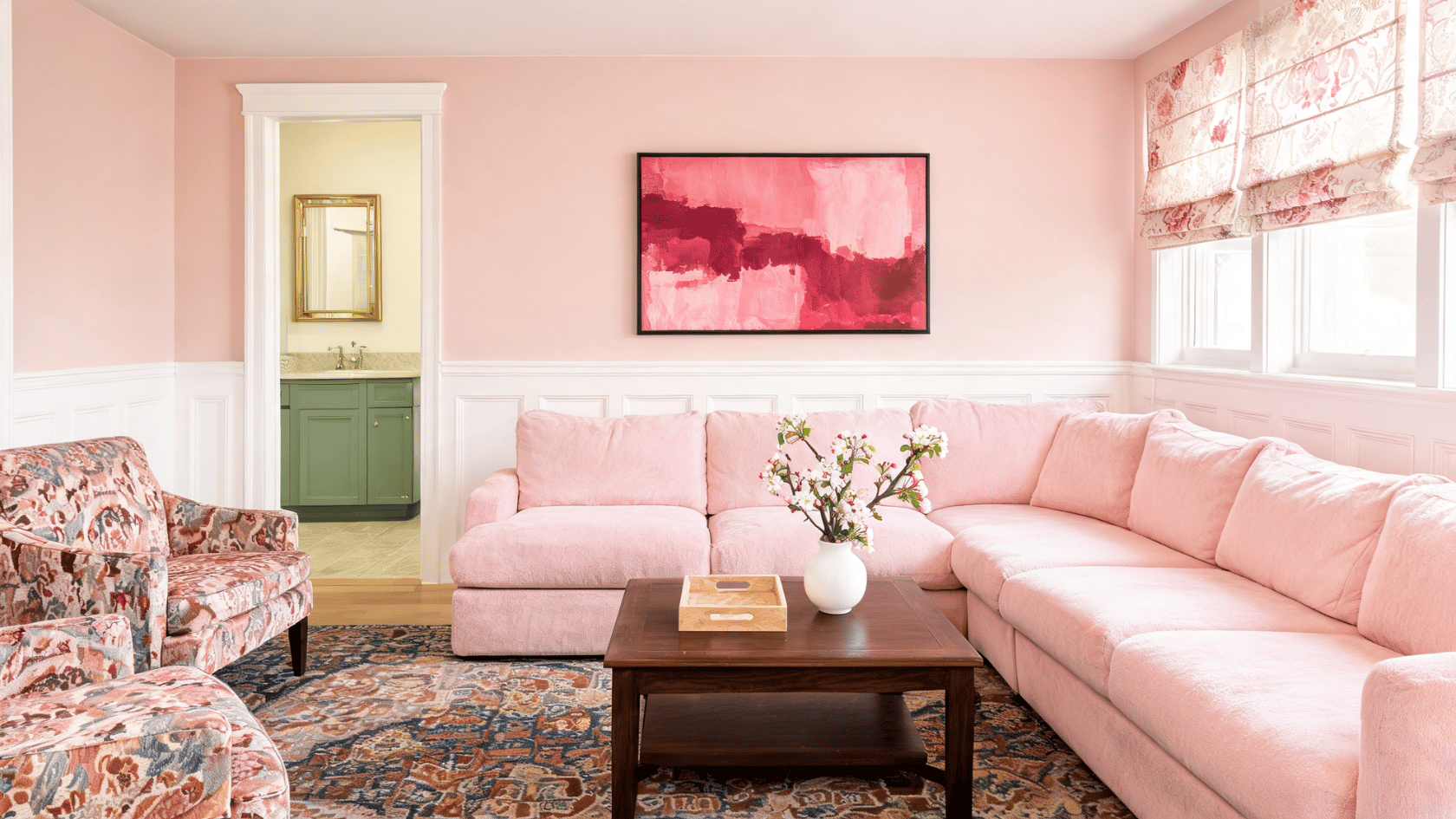 Pink living room with a large sectional sofa, floral armchairs, a wooden coffee table, and abstract wall art.