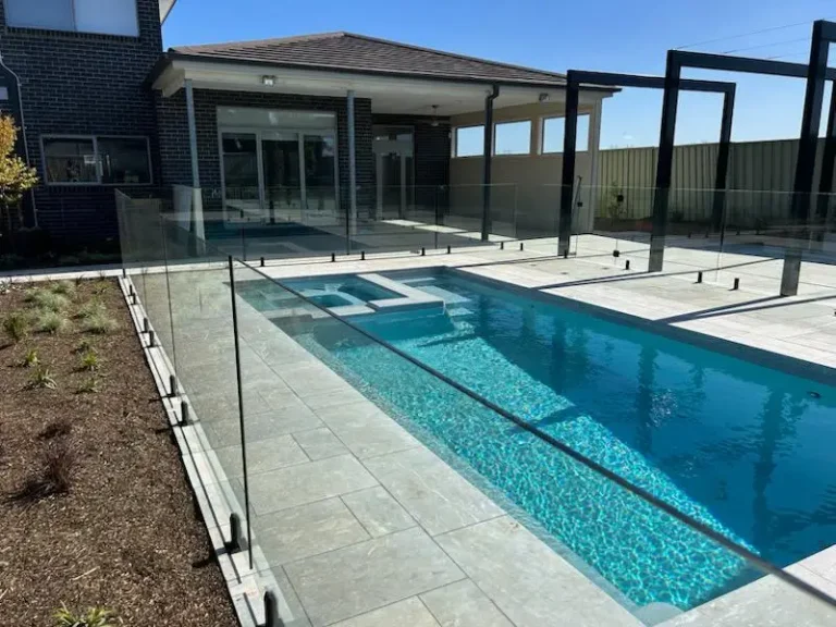 Modern outdoor swimming pool with glass fence and paved poolside area next to residential house