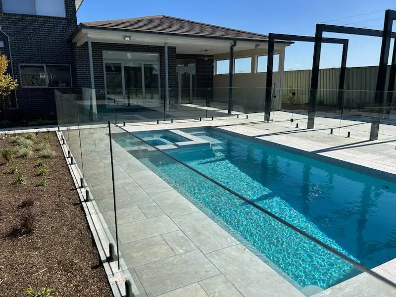 Modern outdoor swimming pool with glass fence and paved poolside area next to residential house