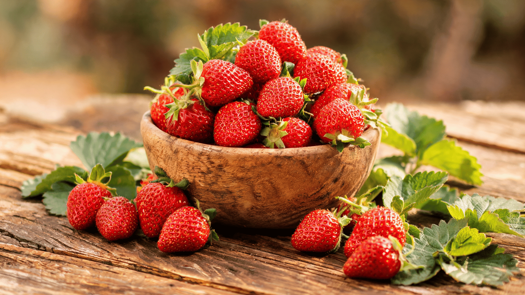 Fresh strawberries in a wooden bowl with strawberry leaves on a rustic surface.