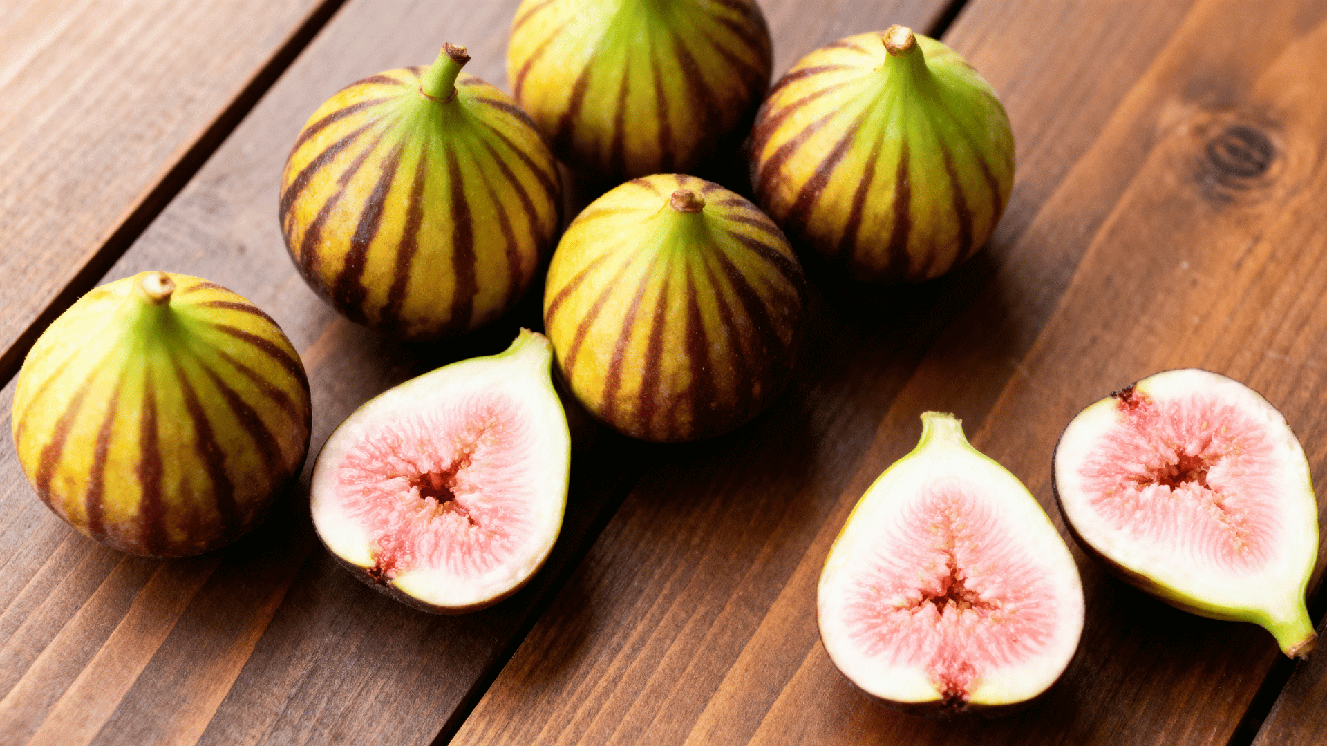 tiger figs with striped green yellow skin placed on wooden table, some cut open showing soft pink interior, soft daylight, natural fresh food photography, high detail, no text no people