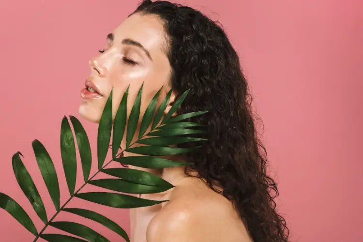 Woman with long curly hair posing with palm leaf against pink background