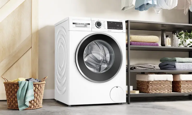 White front-loading washing machine next to laundry basket and shelves with folded towels in modern laundry room