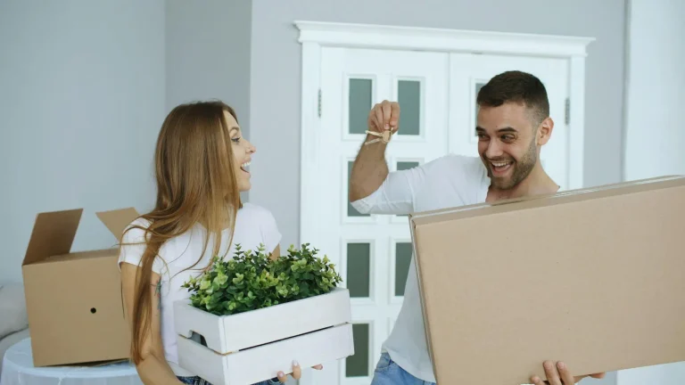 Couple in white shirts holding keys, plant, and moving boxes in bright living room