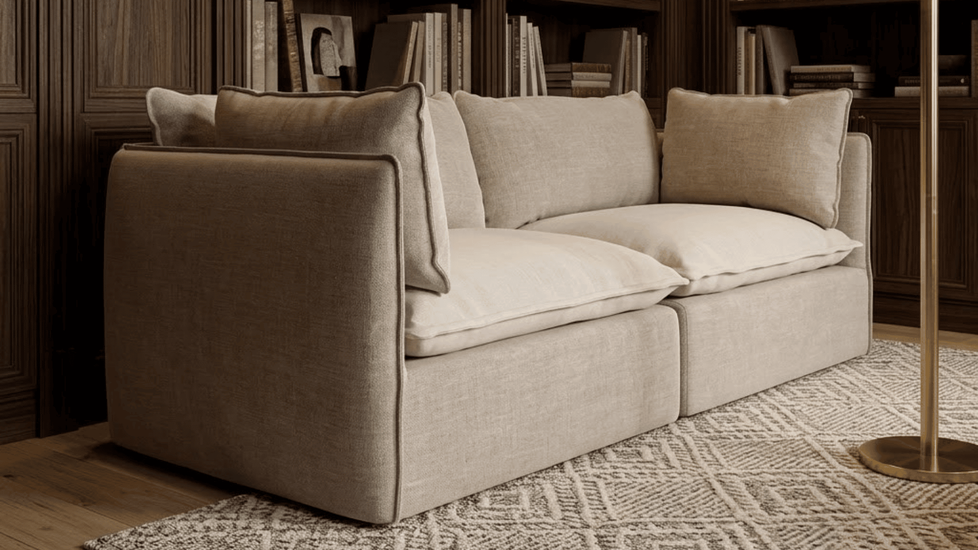 cream two seat sofa with loose cushions beside a brass floor lamp and dark wood bookshelves filled with books