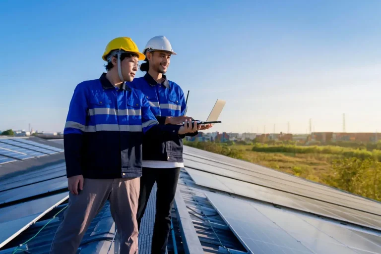 Technicians in blue uniforms inspecting solar panels on rooftop with laptop under clear sky