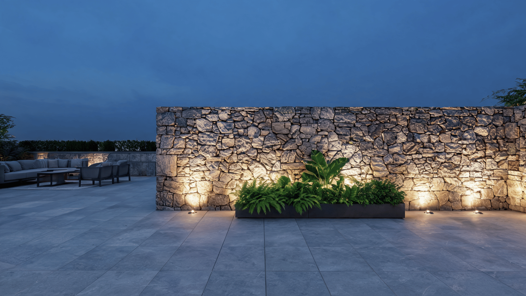 Illuminated faux stone wall paneling in an outdoor patio area at dusk with plants along the base.