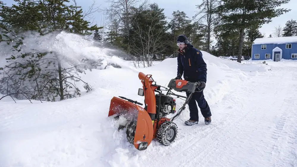 Man using orange snowblower to clear snow near trees and blue house in winter landscape