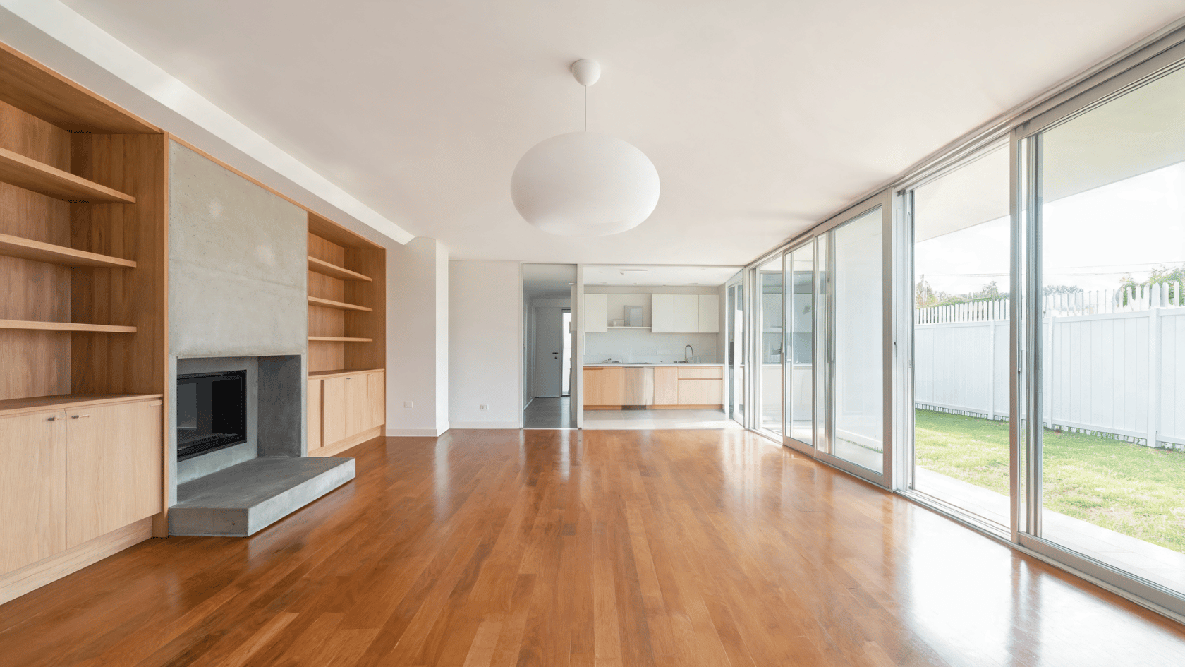 Open living room with glass wall panels, wooden flooring, built-in shelves, fireplace, and kitchen area.