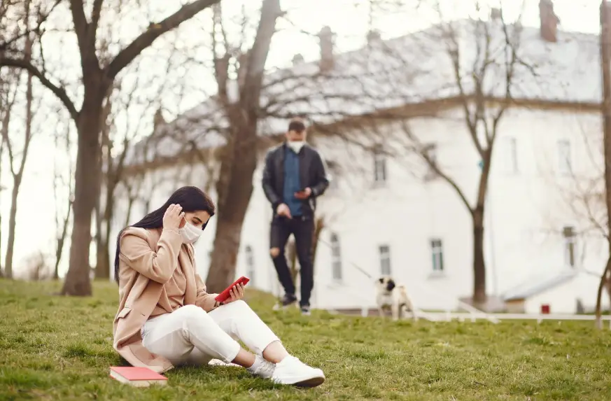 Woman in beige jacket on grass checking phone, man walking dog in background