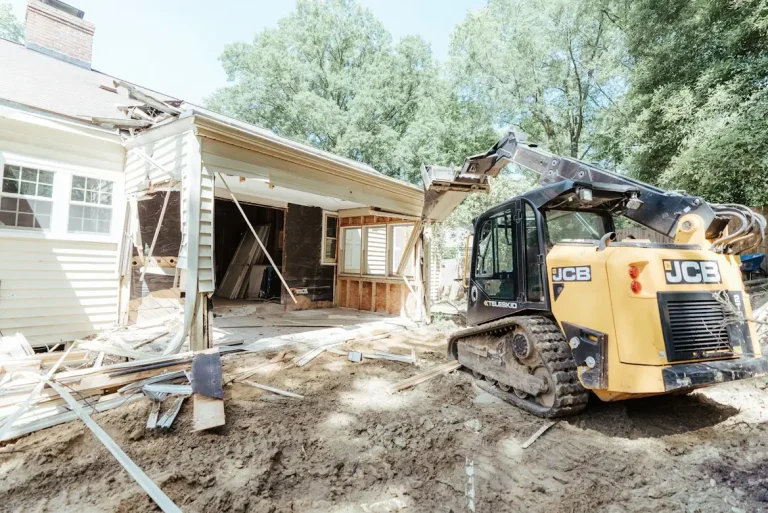 House undergoing demolition with excavator tearing down exterior wall in outdoor setting