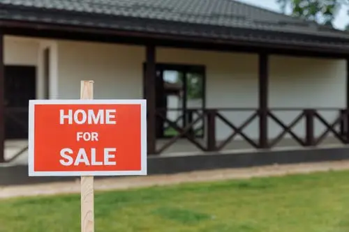 House with a black roof and wooden railings, red "Home for Sale" sign on lawn