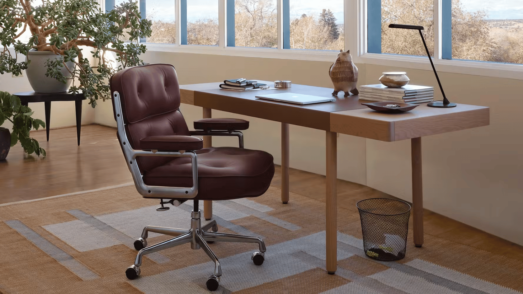Modern home office with wooden desk, leather chair, large windows, indoor plants, and minimalist workspace decor.