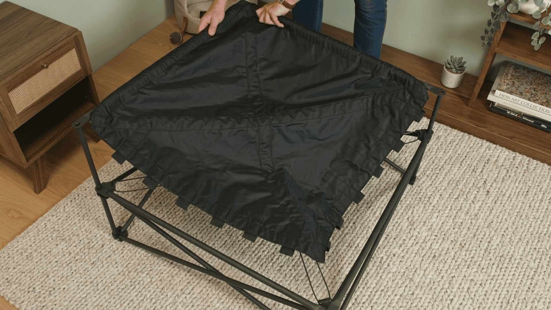 person fitting black fabric support cover over a metal modular sofa frame on a cream rug in a living room